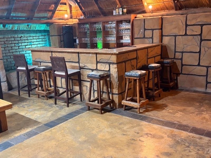 A rustic entertainment bar area featuring a thatched roof, stone-clad bar counter, and built-in glass display cabinetry with concrete flooring.