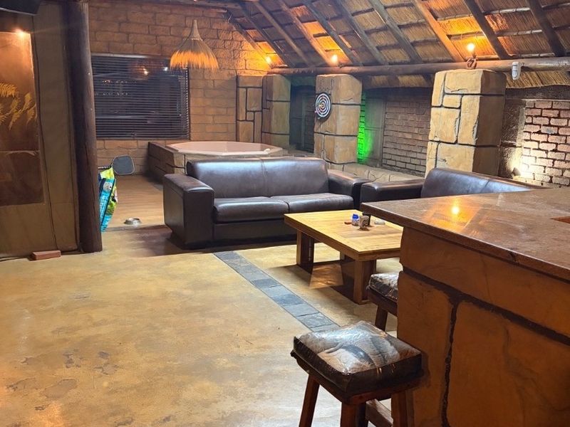 Spacious open-plan entertainment area featuring a rustic thatched roof, exposed timber trusses, stone walls, a built-in bar, and an integrated spa bath.