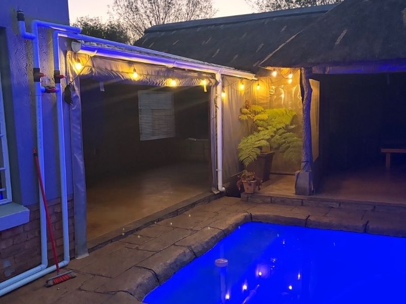 Outdoor pool area featuring a paved deck, covered patio space, and a thatched roof structure, illuminated by ambient string lighting.