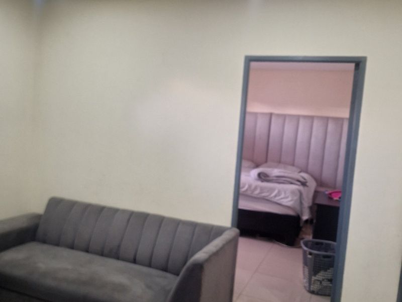 A living room area featuring tiled flooring and recessed downlights, with a doorway leading into an adjacent bedroom.