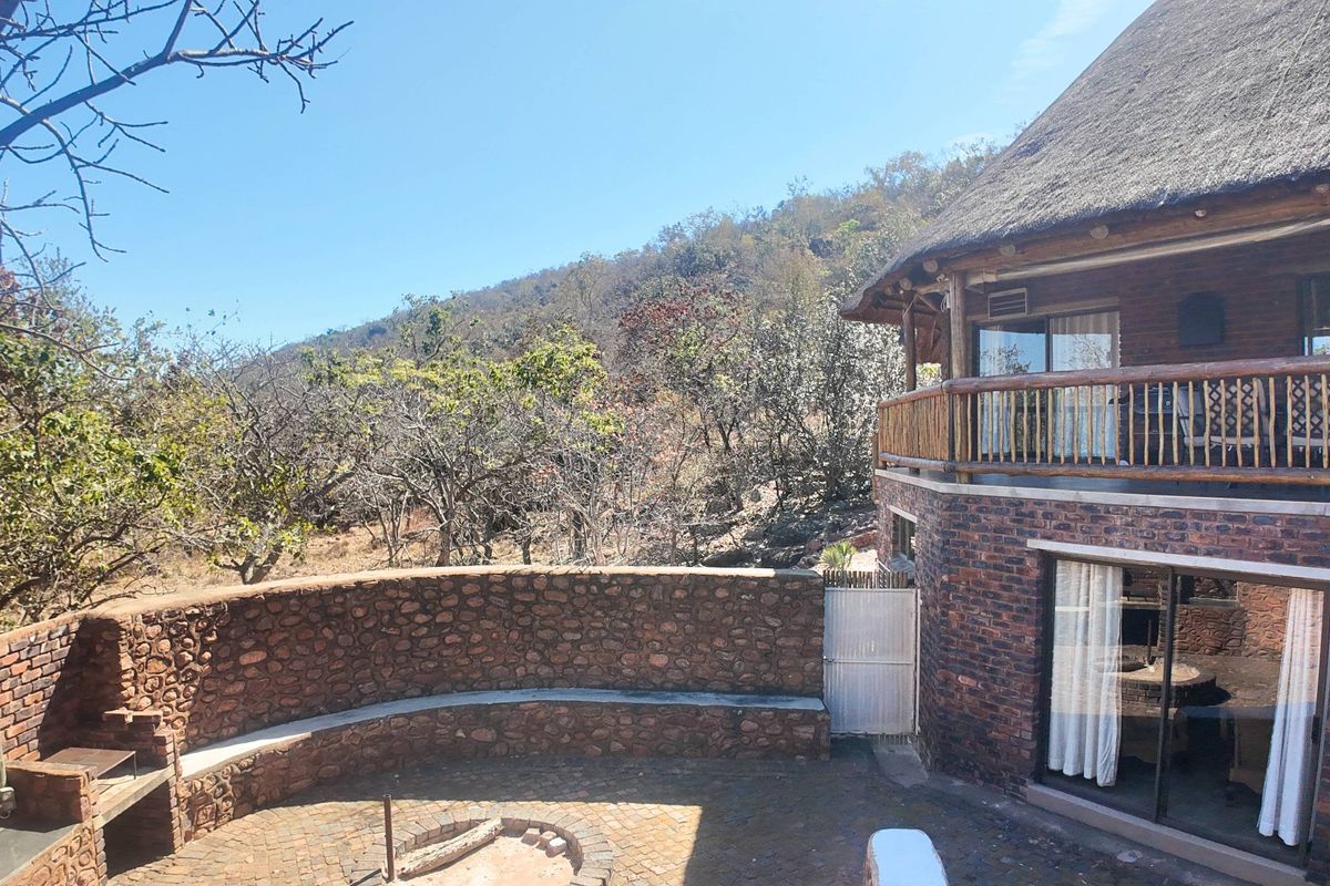 Rear exterior view featuring a thatched roof apartment, brick facade, stone retaining wall with built-in braai, and a paved patio area.