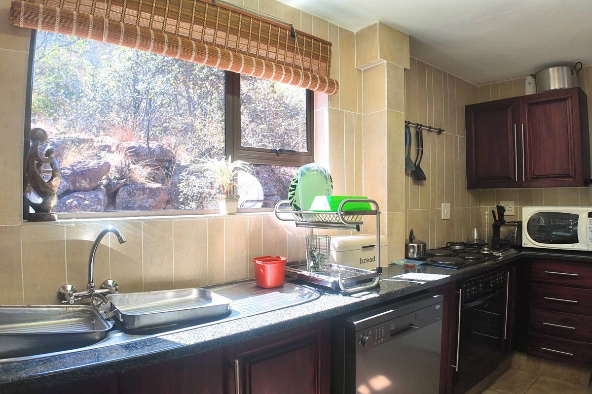 A functional kitchen featuring dark wood cabinetry, granite countertops, a stainless steel sink, and built-in appliances, with a window overlooking a natural rocky landscape.