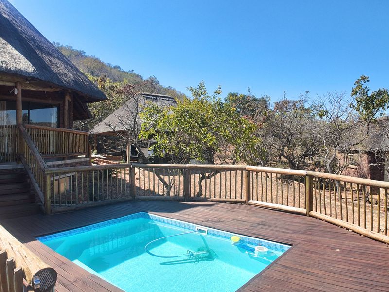 Outdoor pool area featuring timber decking and balustrades, set against a backdrop of thatched-roof structures and natural landscape.