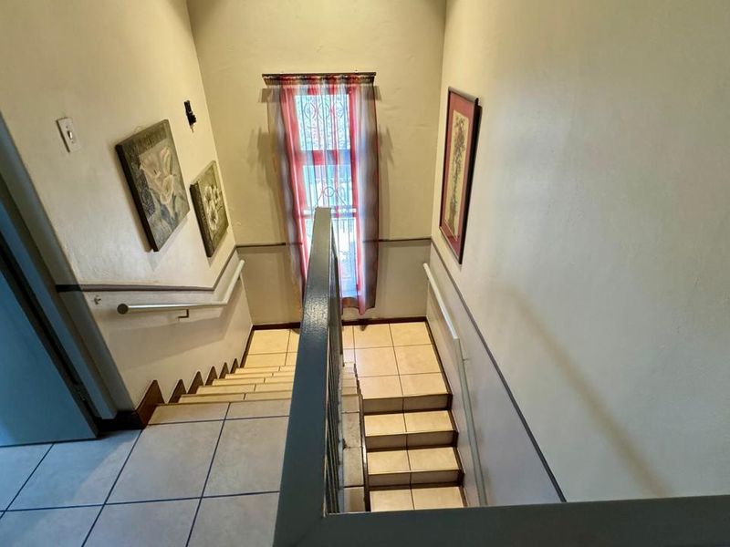 A tiled staircase hallway featuring a metal handrail, chair rail molding, and a window with security bars, providing access between levels.