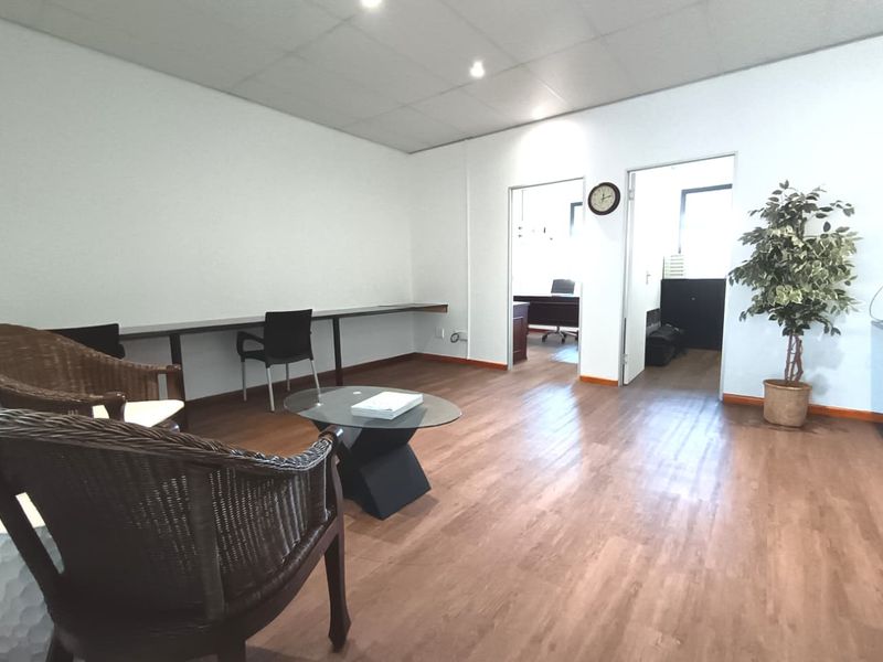 A functional office space featuring laminate flooring, a built-in wall desk, and a suspended ceiling with recessed lighting, providing access to adjacent office rooms.