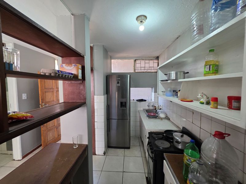 A compact kitchen featuring tiled flooring, a stainless steel sink, built-in open shelving, and a tiled splashback, offering a functional but dated layout.