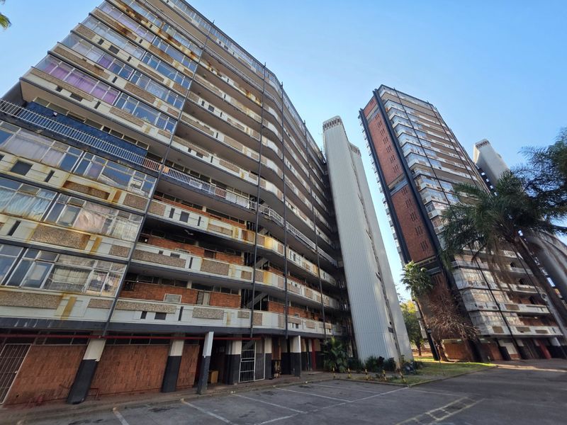 Low-angle view of a large multi-story apartment complex featuring brick and concrete construction with multiple balconies and an adjacent parking area.