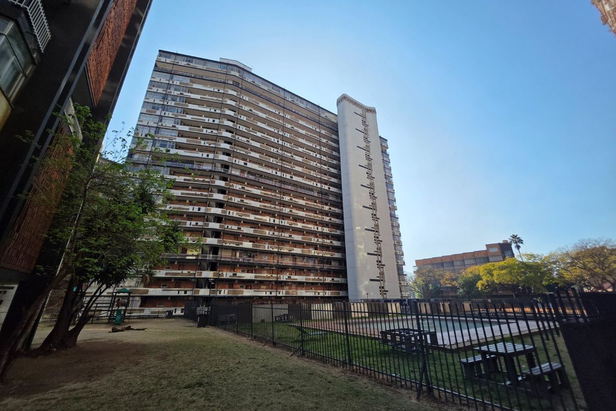 Exterior view of a high-rise apartment complex featuring a communal garden and a fenced swimming pool area.