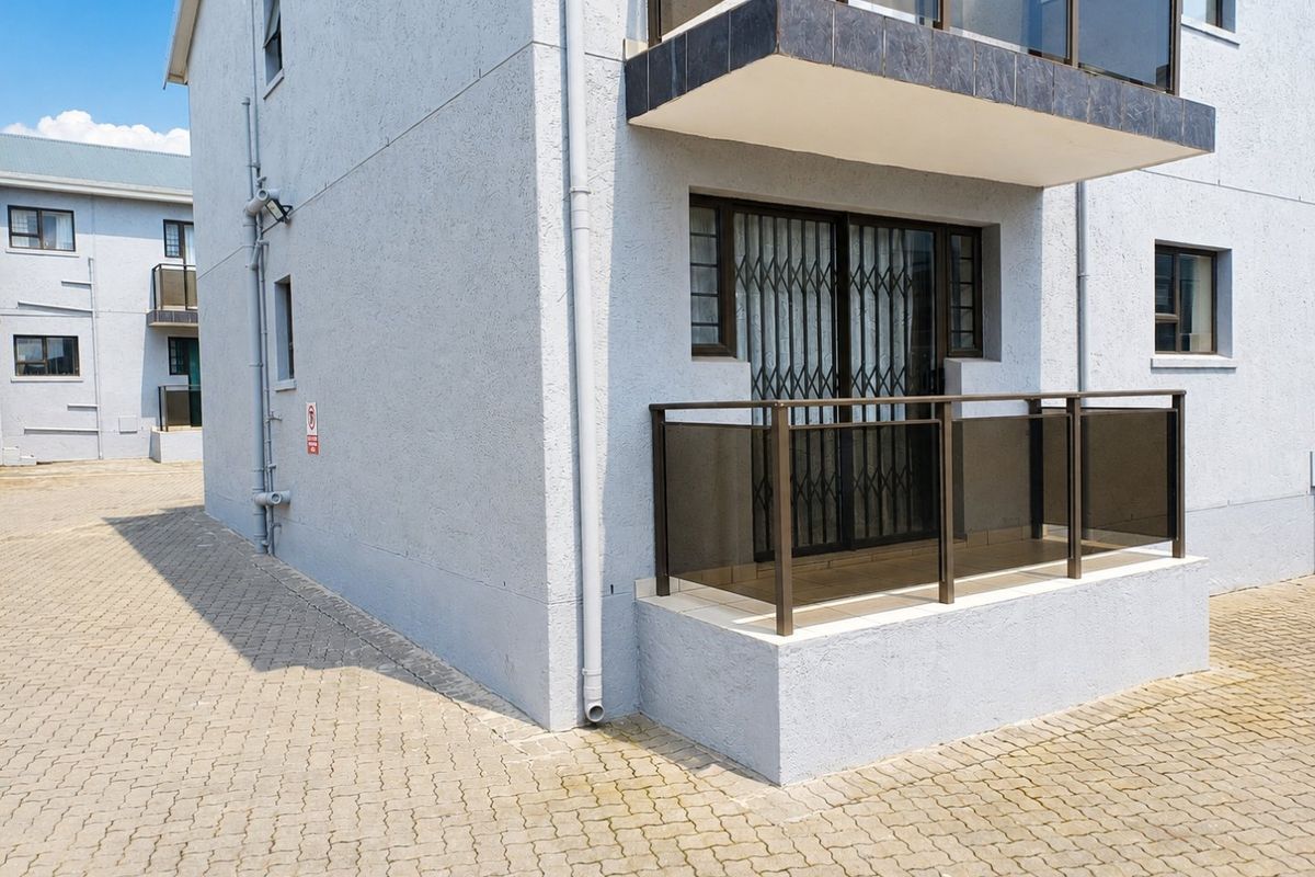 Exterior view of an apartment building featuring a ground-floor balcony with a security gate and paved driveway access.