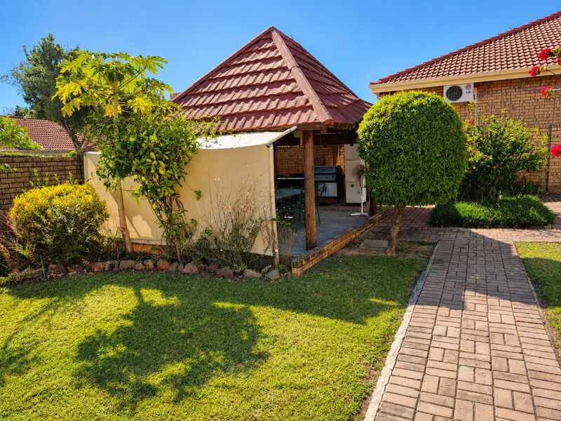 A covered outdoor gazebo area with a tiled roof, situated in a landscaped garden with a paved walkway leading to the main house structure.