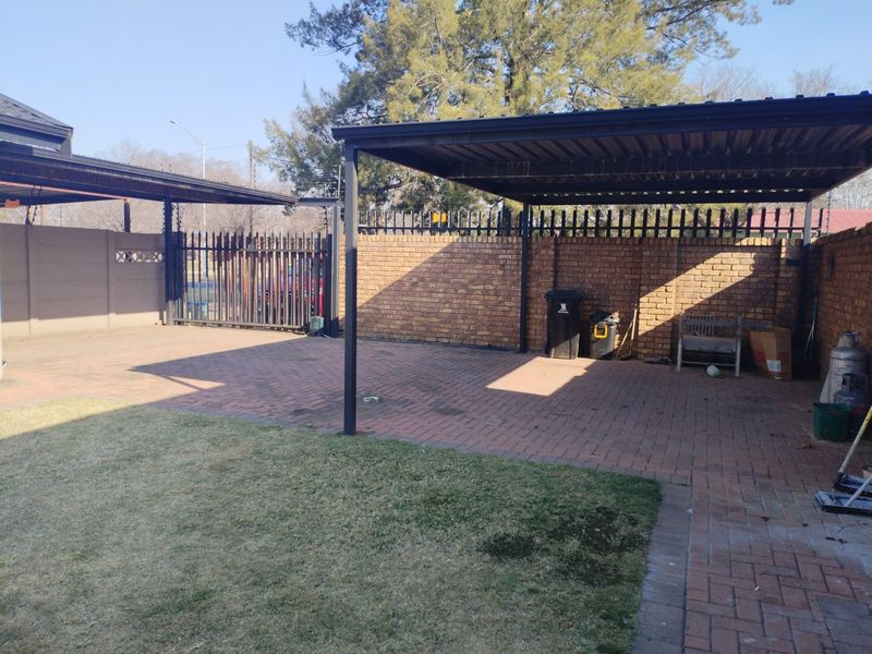 A paved outdoor carport area featuring a metal roof structure, enclosed by brick perimeter walls and secure metal fencing.