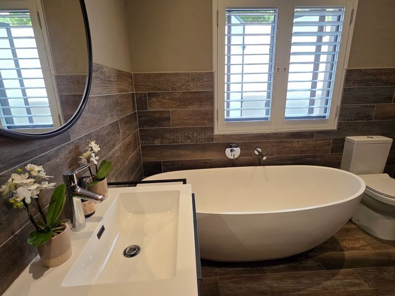 Modern bathroom featuring a sleek freestanding bathtub, wall-mounted basin, and contemporary wood-look tile finishes with plantation-style shutter blinds.