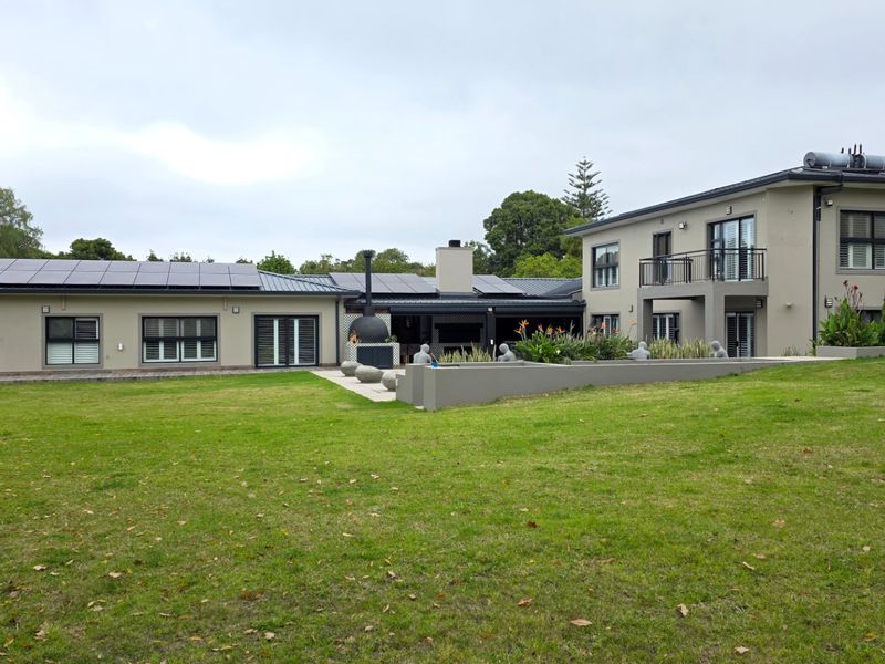 Rear view of a modern two-story house featuring solar panels, a spacious lawn, an outdoor fireplace, and a balcony.