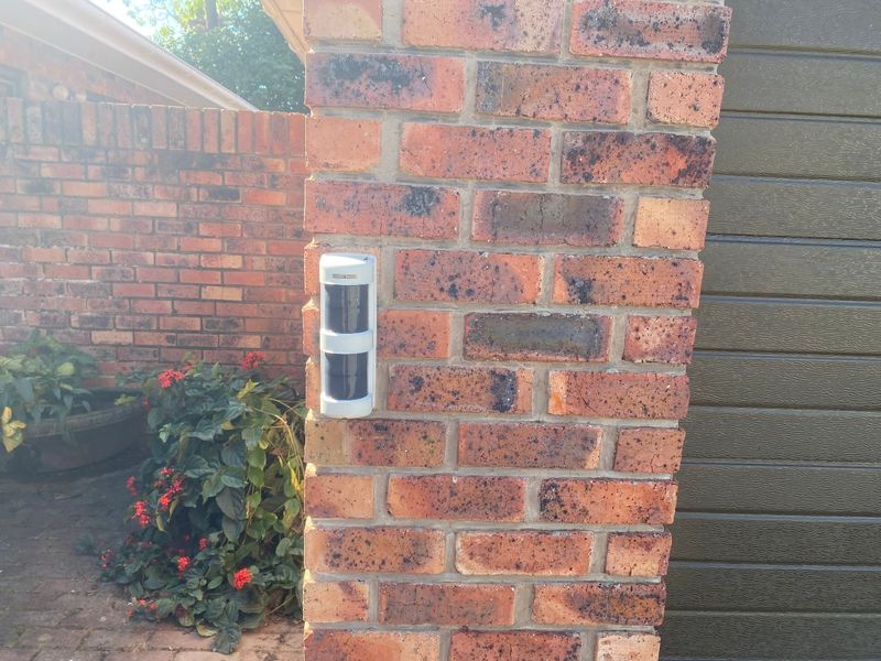 A close-up view of a brick pillar featuring a mounted security sensor, situated near a garage door in an exterior residential setting.