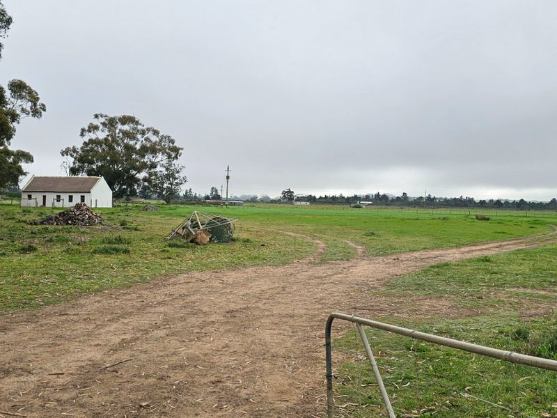 A wide view of a rural property featuring a dirt access track, perimeter fencing, and a small detached outbuilding set in open pasture.
