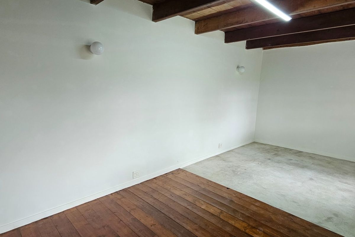 An empty living space featuring exposed timber ceiling beams, a combination of hardwood and concrete flooring, and wall-mounted lighting.