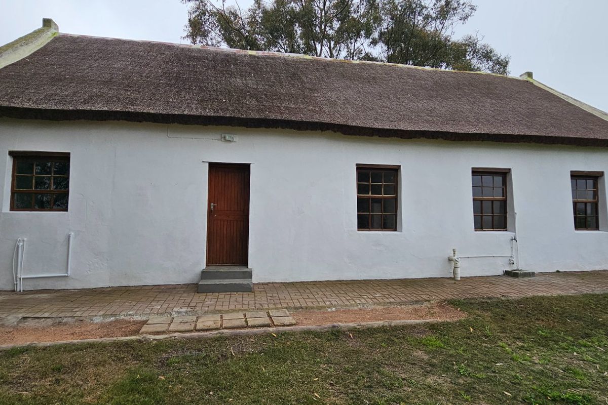 A traditional cottage featuring a thatched roof, white plastered walls, and timber-framed windows, set against a rural landscape with a paved walkway.