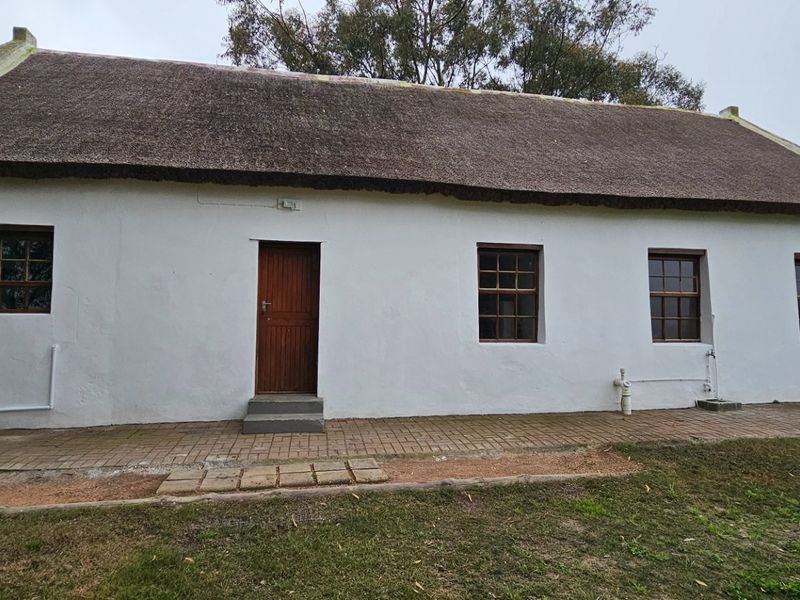 A traditional cottage featuring a thatched roof, white plastered walls, and timber-framed windows, set against a rural landscape with a paved walkway.