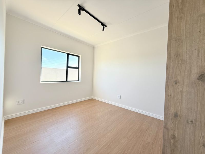A bright, empty bedroom featuring modern laminate flooring, a black aluminium window frame, and contemporary track lighting.