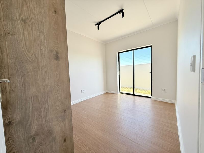 A bright, modern, and empty bedroom featuring light wood-look laminate flooring, track lighting, and a sliding glass door leading to an outdoor area.