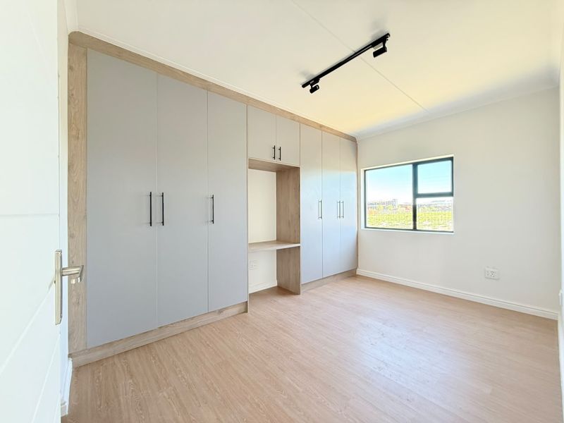 A bright, modern bedroom featuring extensive built-in cupboards with an integrated desk, light wood-look laminate flooring, and contemporary track lighting.