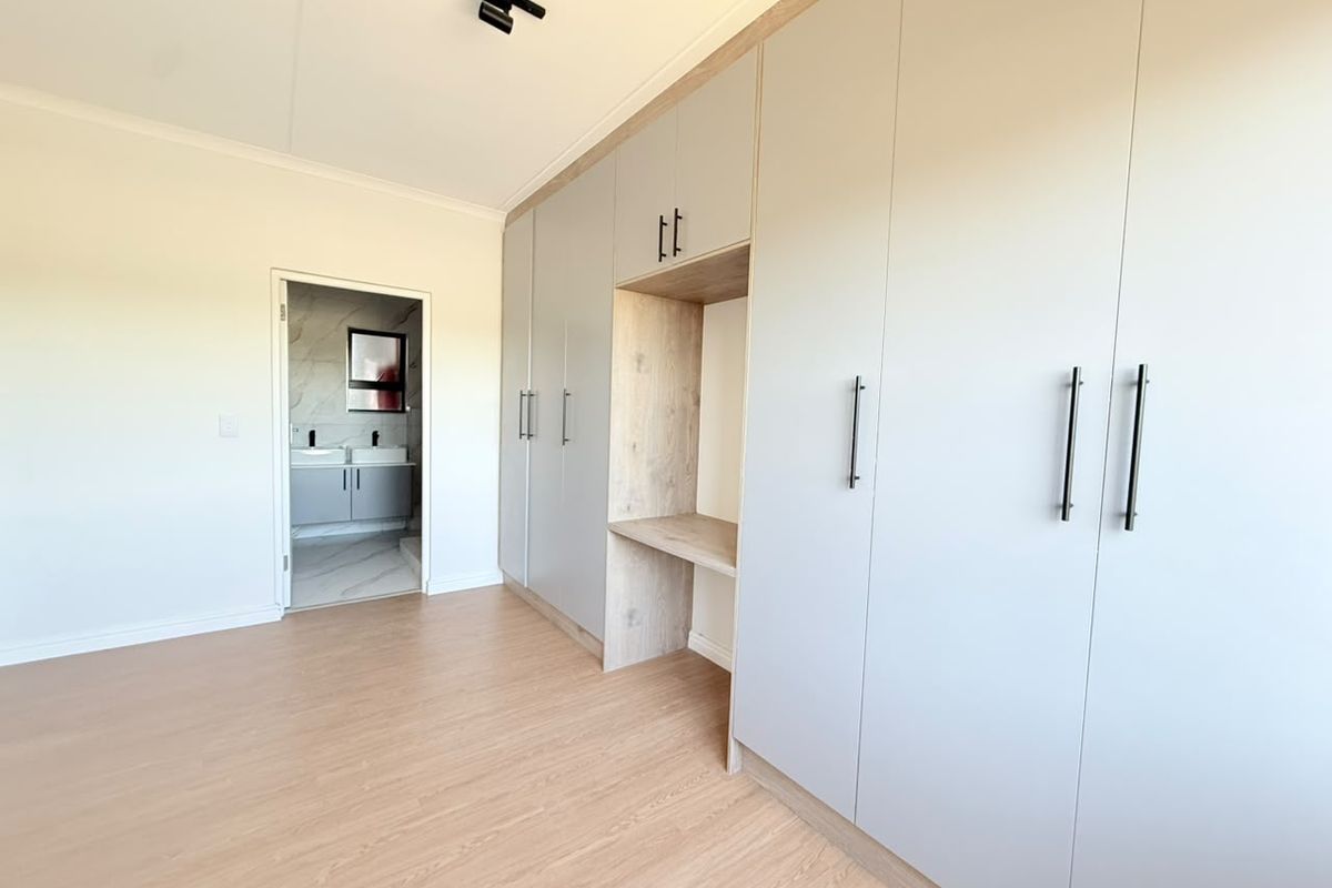 A bright, modern bedroom featuring extensive built-in cupboards with a central vanity nook, light laminate flooring, and direct access to an en-suite bathroom.