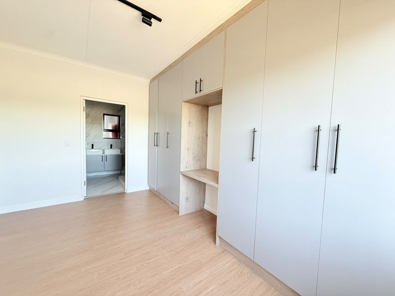 A bright, modern bedroom featuring extensive built-in cupboards with a central vanity nook, light laminate flooring, and direct access to an en-suite bathroom.