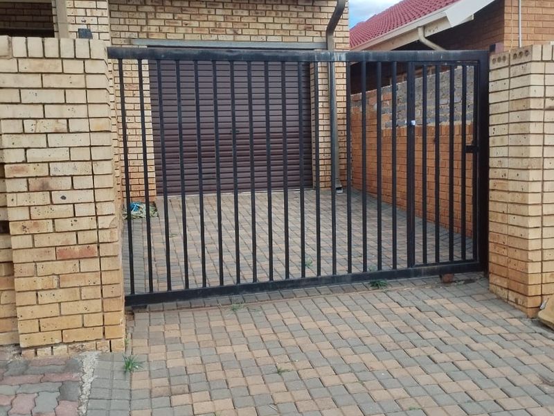 Front view of the property featuring a secure metal sliding gate, paved driveway, and a roller garage door with face-brick exterior walls.