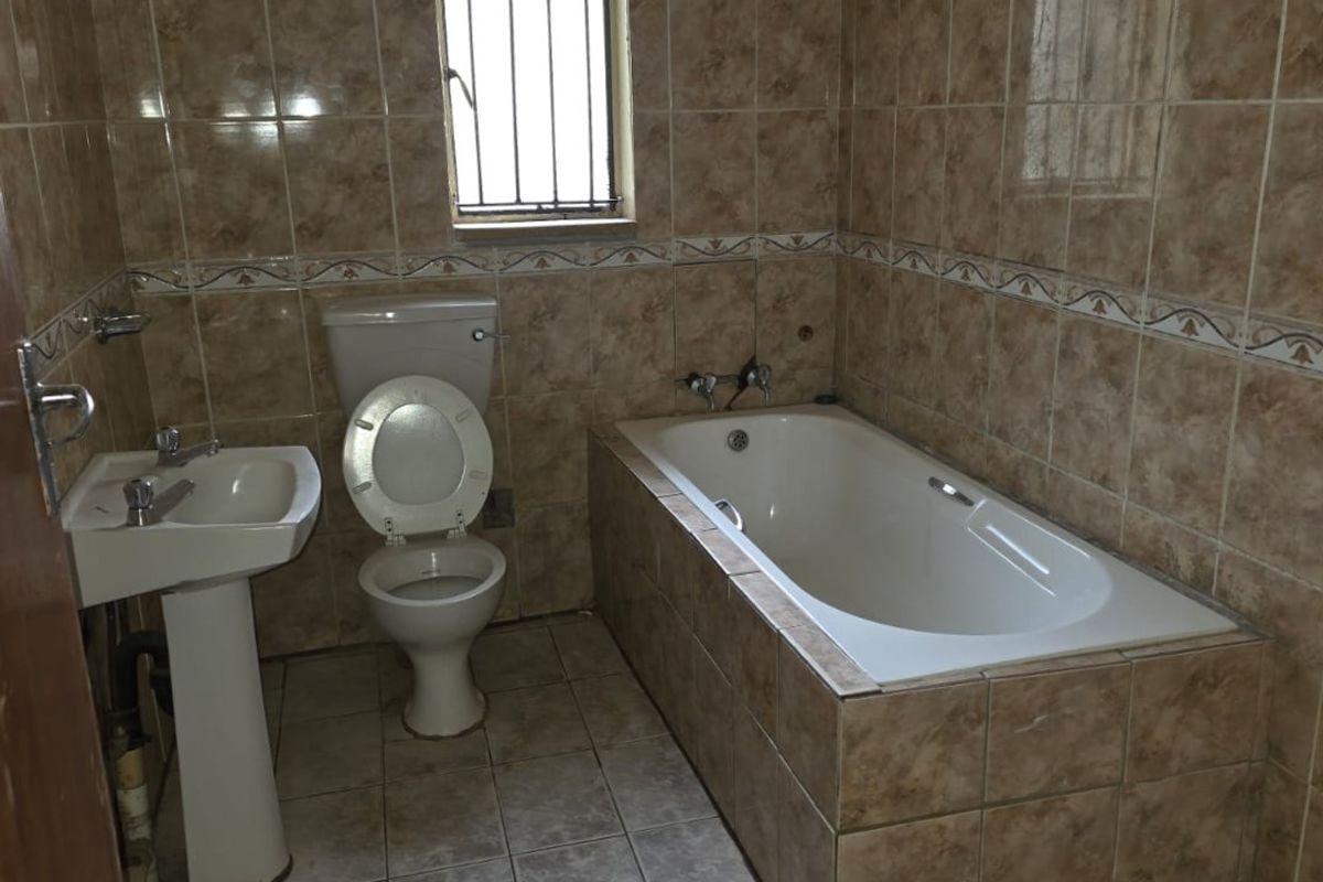 A dated bathroom featuring a pedestal basin, toilet, and built-in bathtub with full-height wall tiling and security bars on the window.