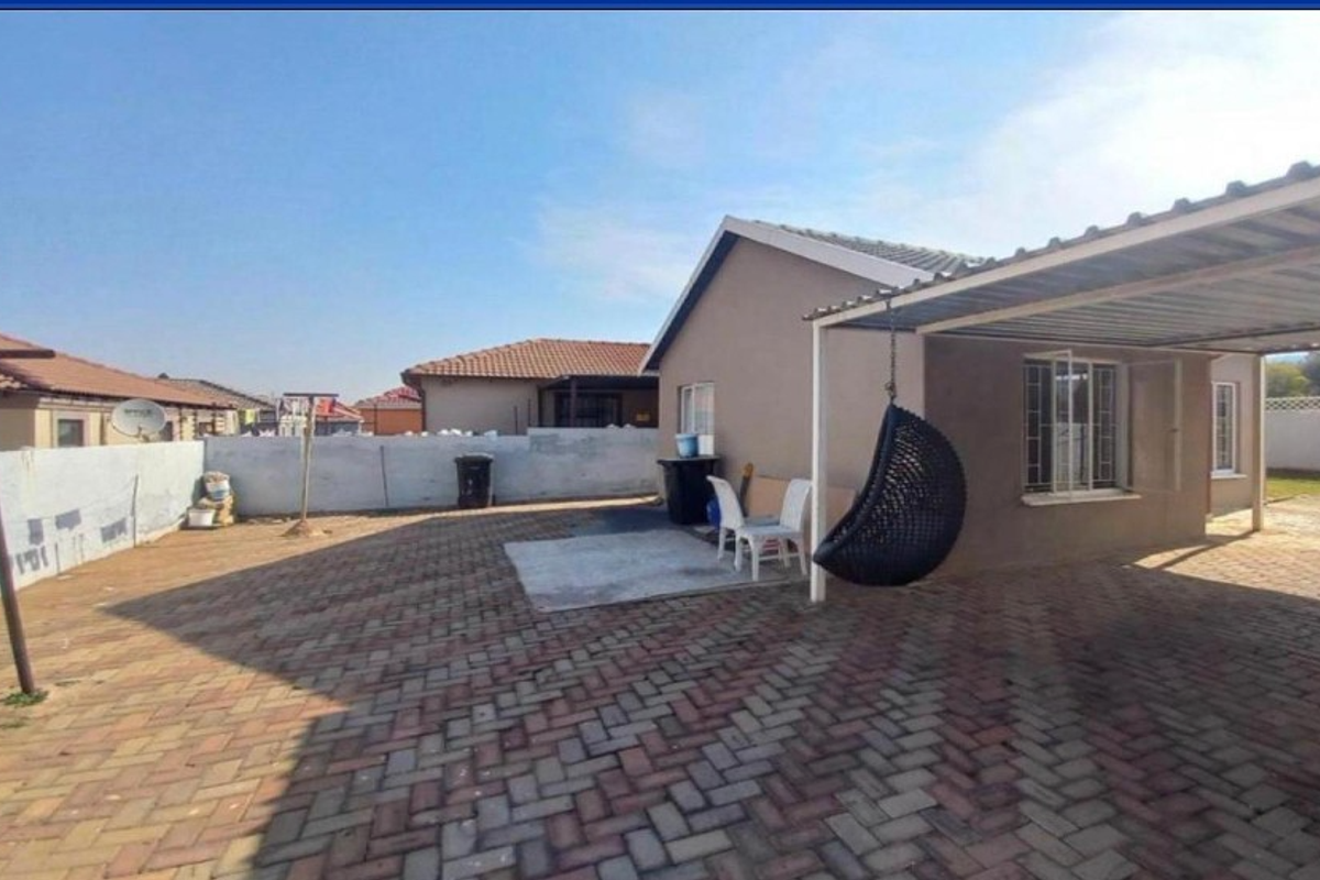 Rear exterior view featuring a paved courtyard, carport, and boundary wall with security bars on the windows.