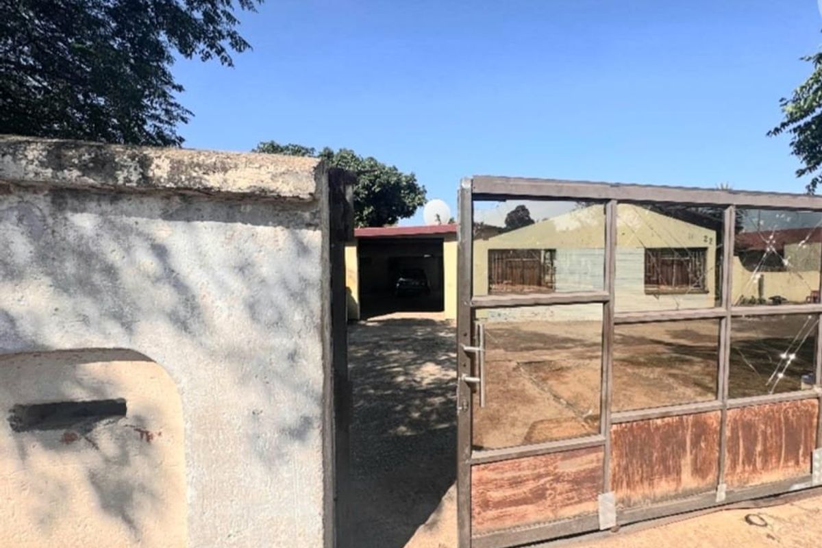 Front entrance view featuring a metal driveway gate, concrete boundary wall, and a carport leading to the main house structure.