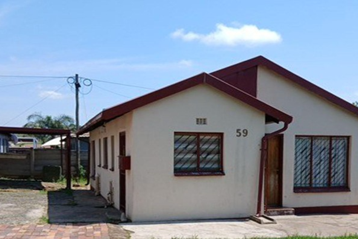 Front view of a single-story residential house featuring a gabled roof, security bars on windows, and an attached carport with a paved driveway.