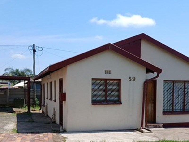 Front view of a single-story residential house featuring a gabled roof, security bars on windows, and an attached carport with a paved driveway.