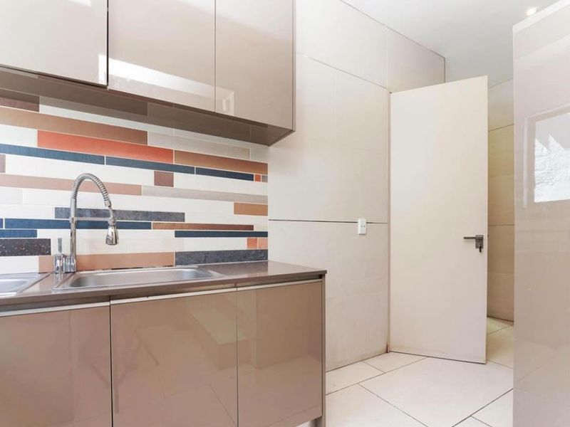 A clean, modern scullery featuring high-gloss cabinetry, a stainless steel sink with a gooseneck faucet, and a decorative multi-colored tiled backsplash.