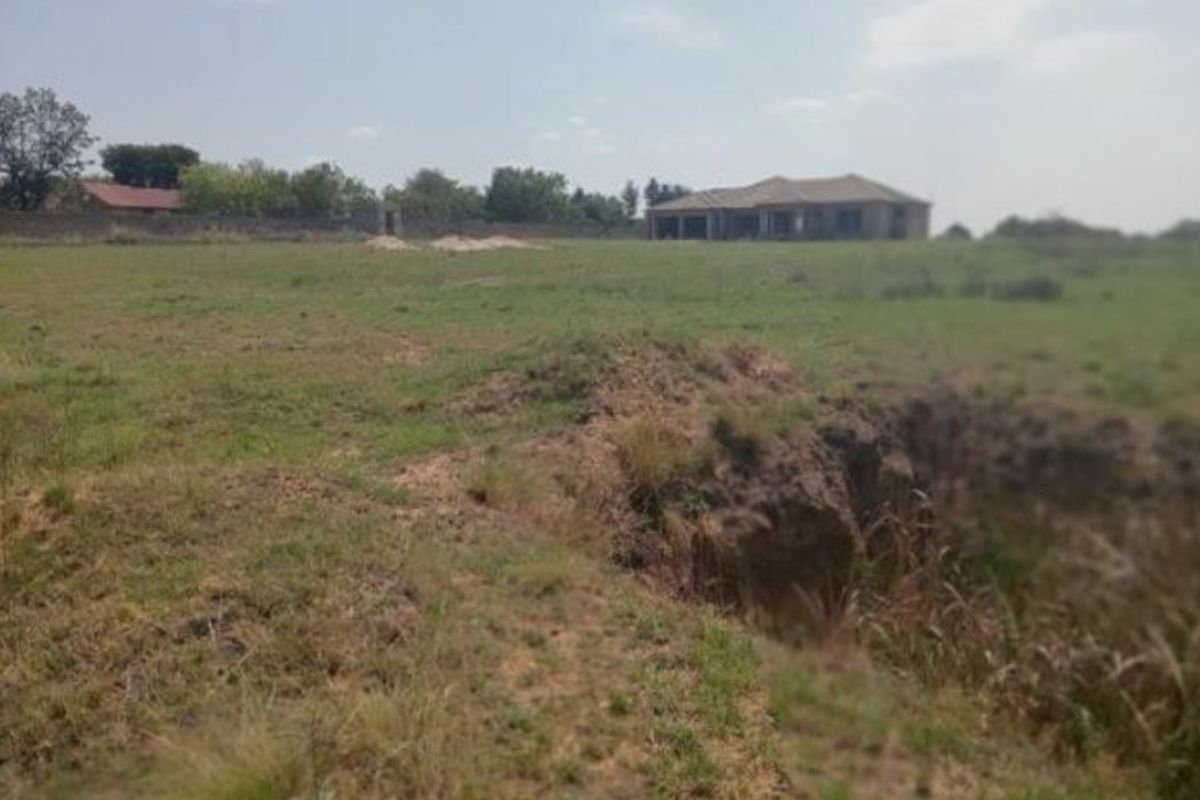 A view of a vacant residential plot featuring uneven grassy terrain, soil erosion, and a boundary wall with a distant structure in the background.