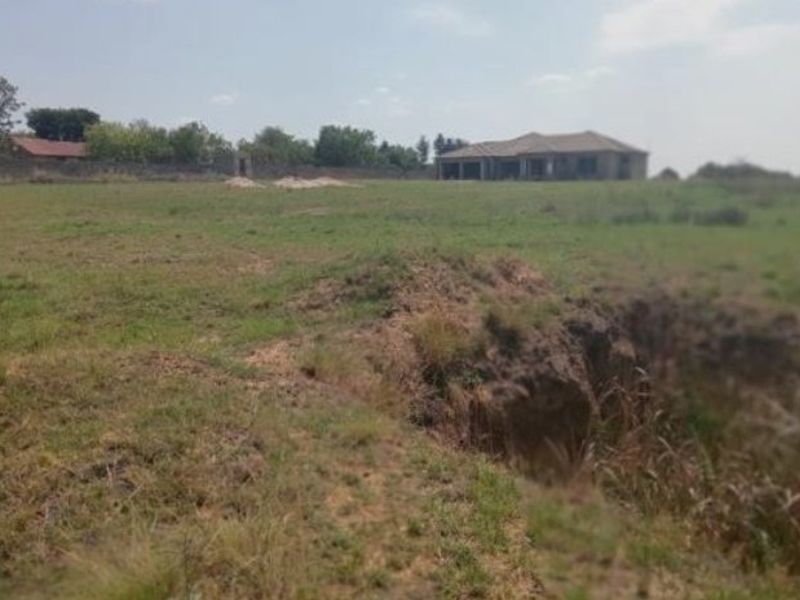 A view of a vacant residential plot featuring uneven grassy terrain, soil erosion, and a boundary wall with a distant structure in the background.