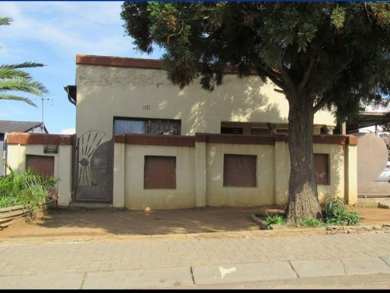 Front elevation of a residential property featuring a boundary wall, metal gate, and mature tree, showing signs of exterior wear and maintenance requirements.