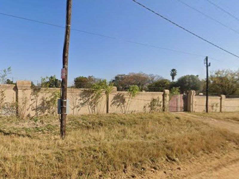 A view of a vacant residential plot featuring a perimeter wall, gated entrance, and utility pole along a dirt road.