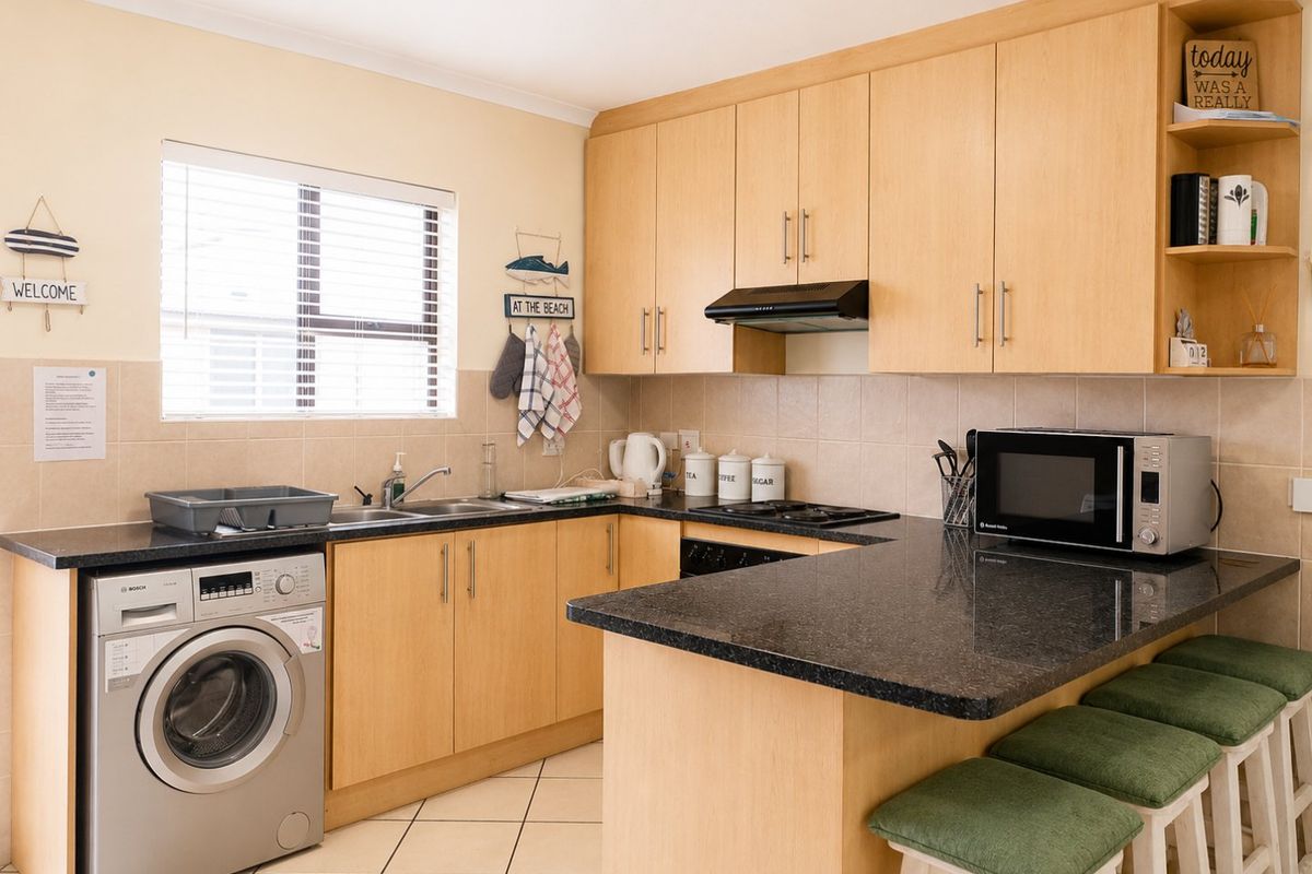 A functional kitchen featuring light wood cabinetry, dark granite countertops, a breakfast bar, and integrated appliances with tiled flooring and backsplash.