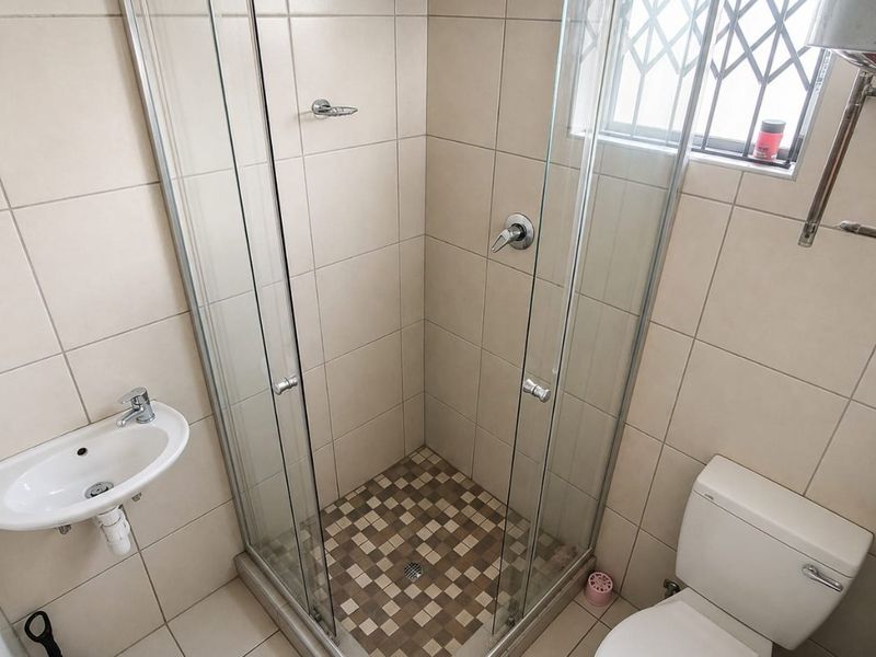 A compact bathroom featuring a glass shower enclosure, wall-mounted basin, and toilet with neutral tiled finishes and a security-grilled window.
