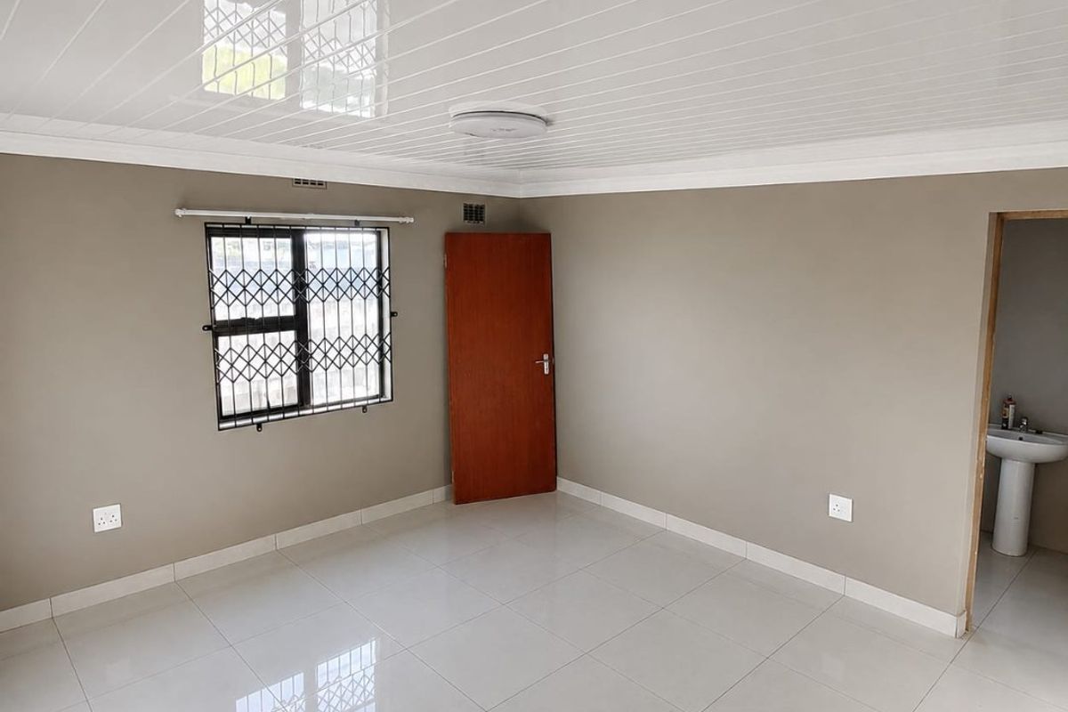 A clean, empty bedroom featuring neutral wall tones, polished tiled flooring, a modern PVC ceiling, and an adjoining en-suite area with a pedestal basin.