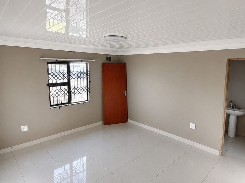 A clean, empty bedroom featuring neutral wall tones, polished tiled flooring, a modern PVC ceiling, and an adjoining en-suite area with a pedestal basin.