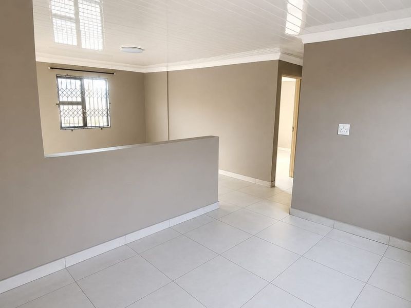 A bright, empty open-plan living area featuring neutral wall tones, white tiled flooring, and a glossy panelled ceiling with integrated lighting.
