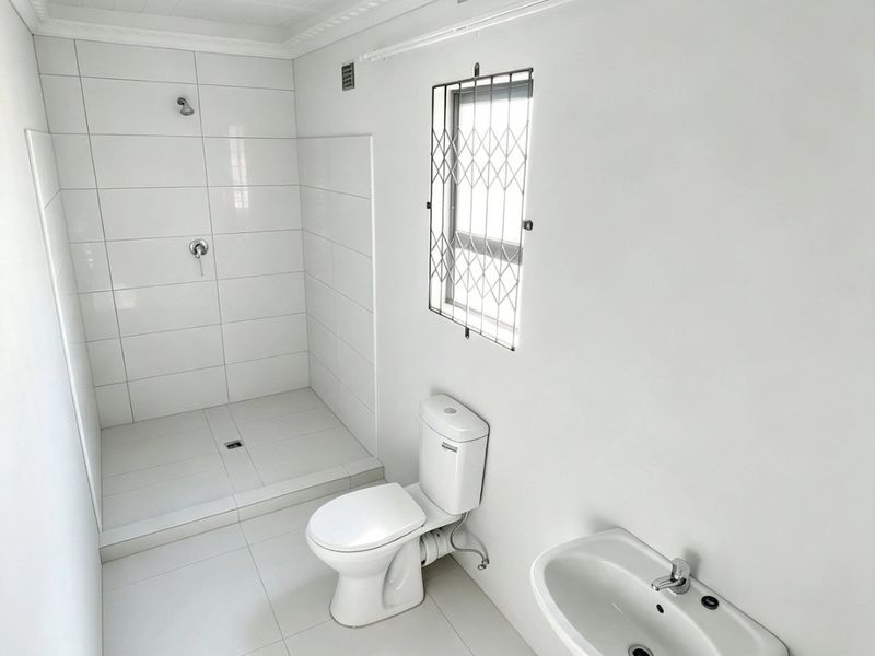 A clean, minimalist bathroom featuring a walk-in shower, pedestal basin, and toilet with white tiled finishes and a secure window.