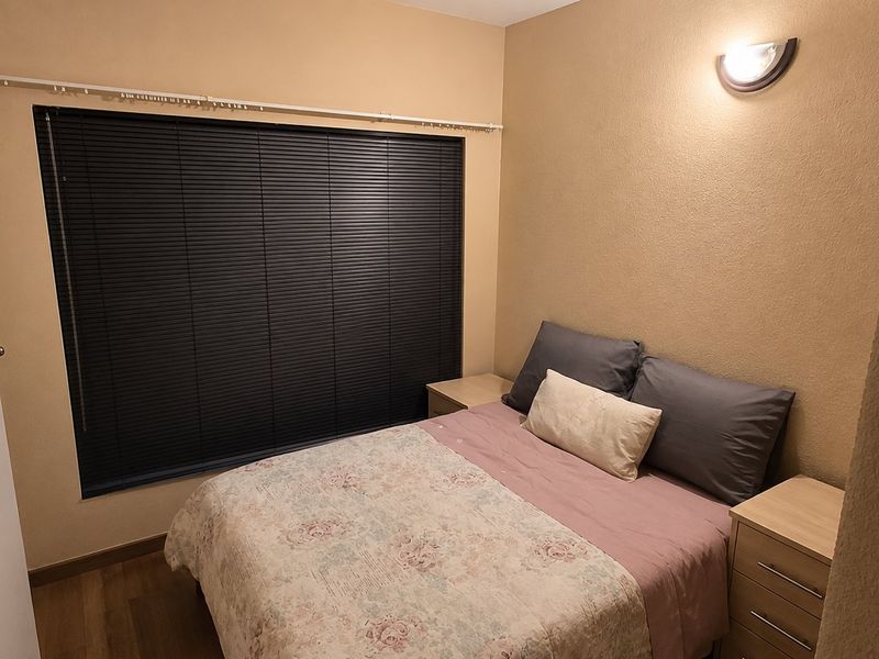 A cozy bedroom featuring laminate flooring, dark venetian blinds, and a wall-mounted light fixture, providing a functional and private sleeping space.