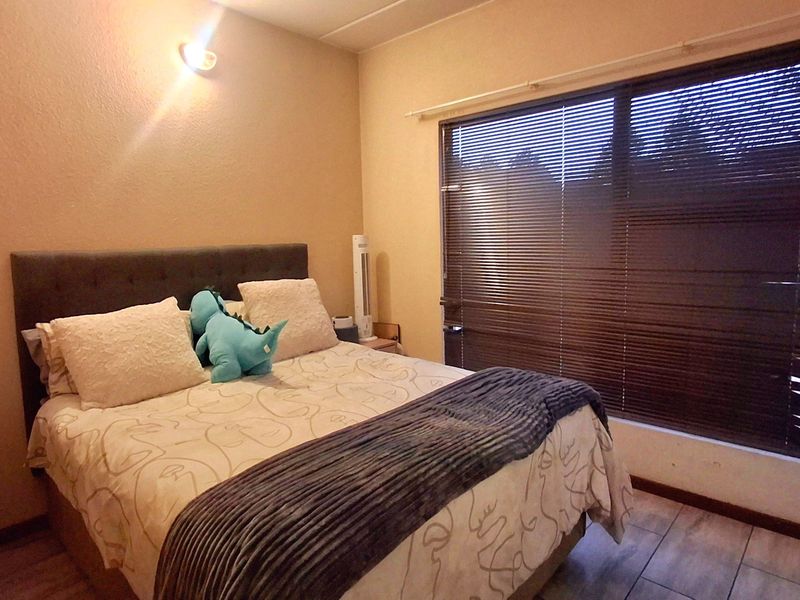 A bedroom featuring laminate flooring, a large window with venetian blinds, and a wall-mounted light fixture.