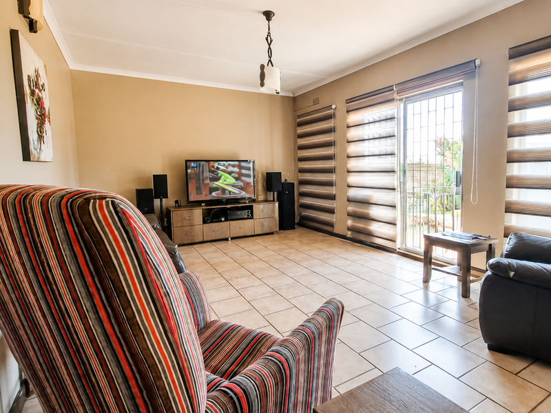 A spacious living room featuring ceramic floor tiles, large windows with roller blinds, and a sliding glass door providing access to the exterior.