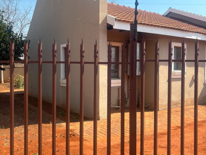 Front view of a single-story house featuring a tiled roof, paved walkway, and secure metal perimeter fencing with electric security wires.