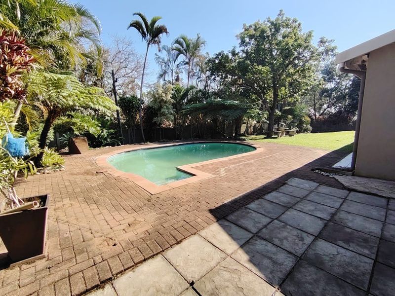 Outdoor pool area featuring a swimming pool, brick and concrete patio space, and a mature landscaped garden.