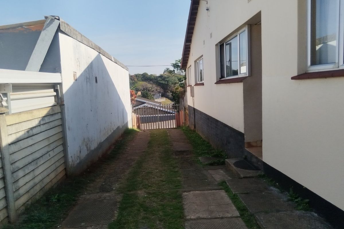 A side exterior view showing a paved driveway leading to a metal security gate, flanked by a residential building and a boundary wall.